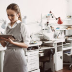 Online production. Close up portrait of young female jeweler in apron using CRM system on her digital tablet Inventory Count